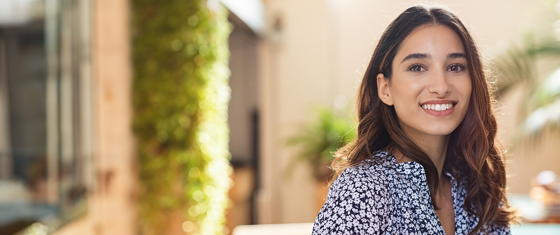 The image features a woman with dark hair smiling at the camera while wearing a patterned top. She stands outdoors against a blurred background, which includes plants and a building.